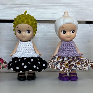 Puede incluir: Cuatro peque&ntilde;as figuras, cada una con un sombrero y un atuendo &uacute;nicos. La primera tiene un sombrero de rat&oacute;n azul y un vestido rosa. La segunda tiene un afro verde y un vestido blanco con lunares negros. La tercera tiene un sombrero de ajo y un vestido morado. La cuarta tiene un sombrero de hipop&oacute;tamo rosa y un vestido verde.