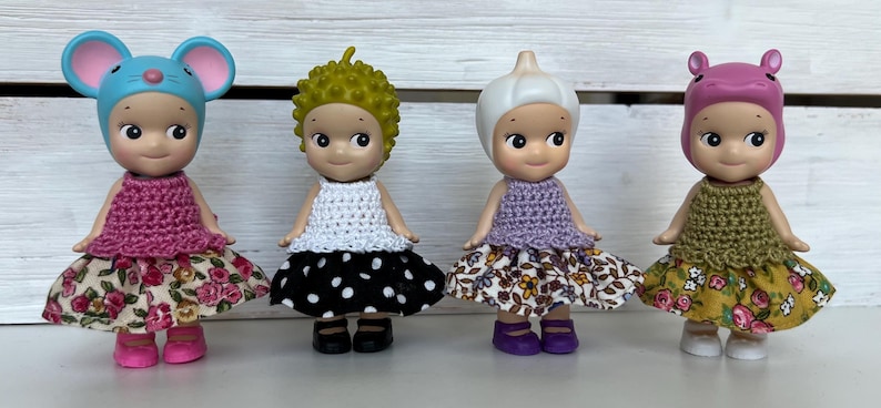 Puede incluir: Cuatro peque&ntilde;as figuras, cada una con un sombrero y un atuendo &uacute;nicos. La primera tiene un sombrero de rat&oacute;n azul y un vestido rosa. La segunda tiene un afro verde y un vestido blanco con lunares negros. La tercera tiene un sombrero de ajo y un vestido morado. La cuarta tiene un sombrero de hipop&oacute;tamo rosa y un vestido verde.