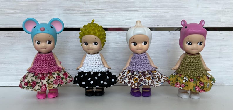 Puede incluir: Cuatro peque&ntilde;as figuras, cada una con un sombrero y un atuendo &uacute;nicos. Las figuras tienen diferentes sombreros: un rat&oacute;n azul, pelo verde puntiagudo, un ajo blanco y un hipop&oacute;tamo rosa. Cada figura lleva un top de ganchillo y una falda floral o de lunares.