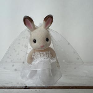 May include: A small, flocked rabbit figurine with brown-tipped ears, wearing a white lace-trimmed dress and a matching sheer white veil featuring a subtle polka dot pattern. The rabbit has black eyes and a pink nose, standing in a formal outfit.