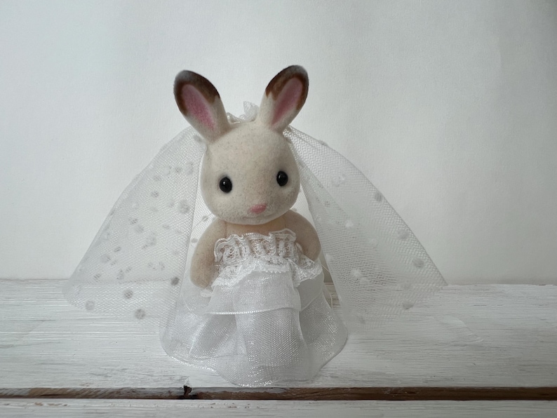 May include: A small, flocked rabbit figurine with brown-tipped ears, wearing a white lace-trimmed dress and a matching sheer white veil featuring a subtle polka dot pattern. The rabbit has black eyes and a pink nose, standing in a formal outfit.