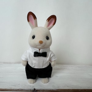 May include: A flocked rabbit figurine with cream-colored fur and brown-tipped ears. The rabbit is dressed in a white collared shirt with a small black bow tie and black trousers. The figure has large black eyes and a small tan nose.