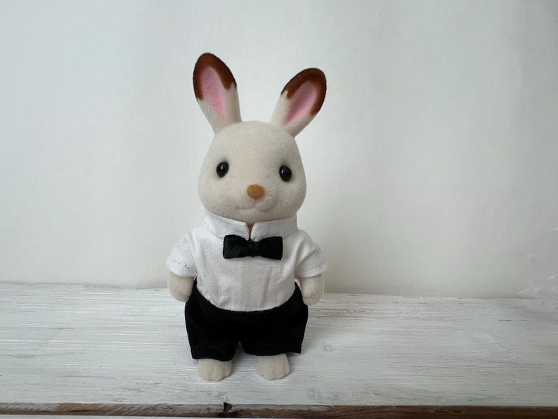 May include: A flocked rabbit figurine with cream-colored fur and brown-tipped ears. The rabbit is dressed in a white collared shirt with a small black bow tie and black trousers. The figure has large black eyes and a small tan nose.