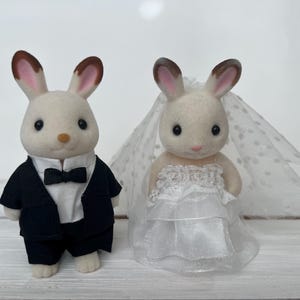 May include: Two flocked rabbit figurines. The left figure wears a black tuxedo jacket, white shirt, and black bow tie. The right figure wears a white tiered wedding dress with lace detailing and a matching white polka-dot tulle veil over its ears.