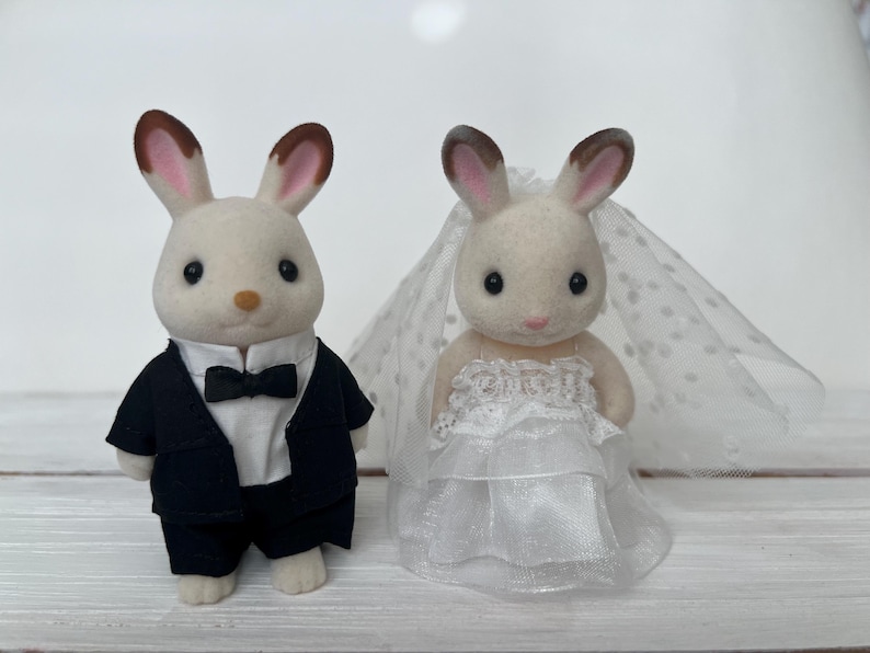 May include: Two flocked rabbit figurines. The left figure wears a black tuxedo jacket, white shirt, and black bow tie. The right figure wears a white tiered wedding dress with lace detailing and a matching white polka-dot tulle veil over its ears.