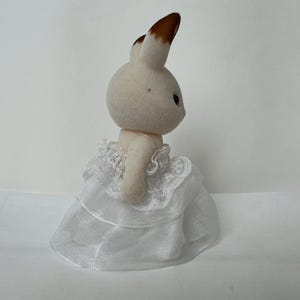 May include: A small, flocked rabbit figurine with cream-colored fur and brown-tipped ears. The rabbit is wearing a white, multi-layered dress made of sheer, translucent fabric with delicate lace trim detailing around the bodice and skirt hem.