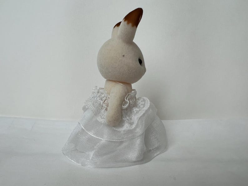 May include: A small, flocked rabbit figurine with cream-colored fur and brown-tipped ears. The rabbit is wearing a white, multi-layered dress made of sheer, translucent fabric with delicate lace trim detailing around the bodice and skirt hem.