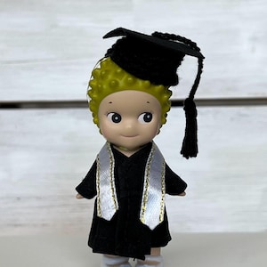 May include: A small figurine of a graduate wearing a black cap and gown with a white and gold sash. The figurine has a light skin tone, green hair, and is wearing white shoes. The graduation cap has a black tassel.