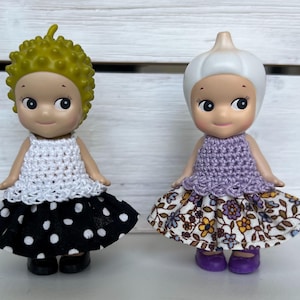 Puede incluir: Dos peque&ntilde;as figuras. Una tiene cabello amarillo, una parte superior de ganchillo blanca y una falda de lunares en blanco y negro. La otra tiene una cabeza blanca en forma de bulbo, una parte superior de ganchillo p&uacute;rpura y una falda floral.