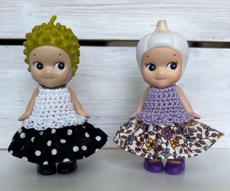 Puede incluir: Dos peque&ntilde;as figuras. Una tiene cabello amarillo, una parte superior de ganchillo blanca y una falda de lunares en blanco y negro. La otra tiene una cabeza blanca en forma de bulbo, una parte superior de ganchillo p&uacute;rpura y una falda floral.