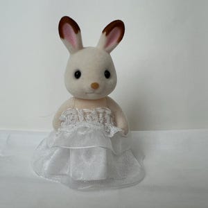 May include: A flocked rabbit figurine with cream-colored fur and brown-tipped ears. The rabbit is wearing a white dress featuring a layered, ruffled skirt made of sheer fabric and a delicate lace trim detail across the bodice area.
