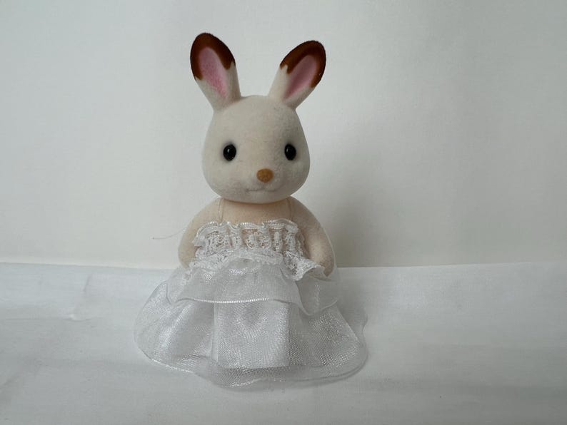 May include: A flocked rabbit figurine with cream-colored fur and brown-tipped ears. The rabbit is wearing a white dress featuring a layered, ruffled skirt made of sheer fabric and a delicate lace trim detail across the bodice area.
