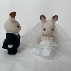 May include: Two flocked rabbit figurines dressed for a wedding. One rabbit stands in profile wearing a dark navy blue suit with a white shirt. The other rabbit faces forward wearing a white layered dress and a sheer white veil with polka dots.