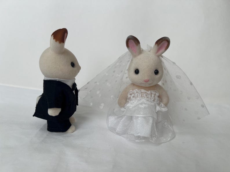 May include: Two flocked rabbit figurines dressed for a wedding. One rabbit stands in profile wearing a dark navy blue suit with a white shirt. The other rabbit faces forward wearing a white layered dress and a sheer white veil with polka dots.
