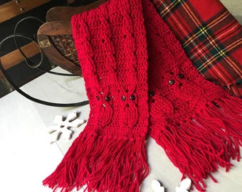 May include: A bright red crocheted scarf featuring a cable knit pattern and owl motifs with dark button eyes. The scarf has long, thick fringe at both ends and is constructed from yarn, showcasing a textured design with multiple owl figures.