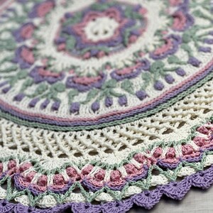 Pu&ograve; includere: Un centrino rotondo all'uncinetto con un complesso motivo a mandala in filato color crema, verde salvia, rosa antico e lavanda. Il design presenta intricati motivi floreali, punti a rete traforati e un bordo smerlato con finitura viola.