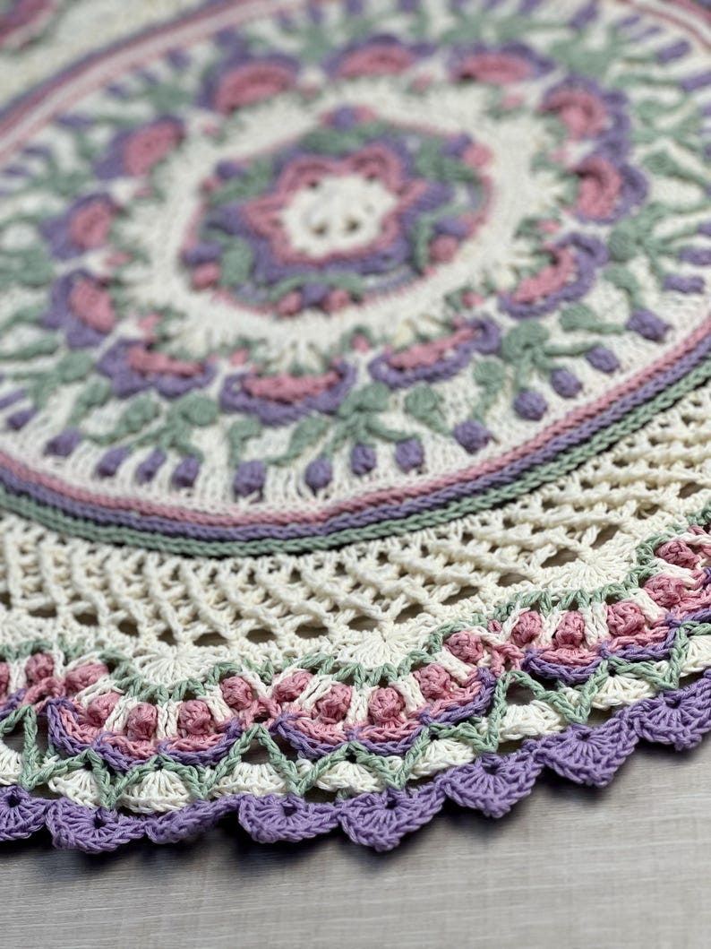Pu&ograve; includere: Un centrino rotondo all'uncinetto con un complesso motivo a mandala in filato color crema, verde salvia, rosa antico e lavanda. Il design presenta intricati motivi floreali, punti a rete traforati e un bordo smerlato con finitura viola.