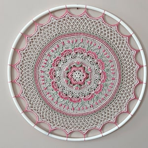 Pu&ograve; includere: Una decorazione da parete circolare con un intricato lavoro all'uncinetto nei toni del crema, verde salvia e rosa antico. Il design &egrave; centrato su un cerchio bianco, con un complesso motivo a mandala che si irradia dal centro.