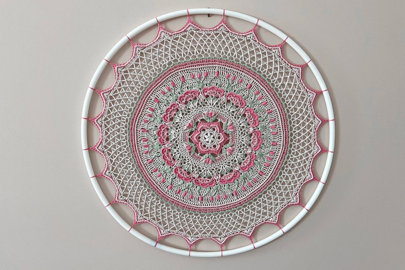 Pu&ograve; includere: Una decorazione da parete circolare con un intricato lavoro all'uncinetto nei toni del crema, verde salvia e rosa antico. Il design &egrave; centrato su un cerchio bianco, con un complesso motivo a mandala che si irradia dal centro.