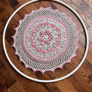 Pu&ograve; includere: Un mandala circolare all'uncinetto con intricati motivi floreali nelle tonalit&agrave; crema, rosa tenue e verde salvia. Il pezzo &egrave; montato all'interno di un grande cerchio in plastica bianca, che mette in mostra un design dettagliato con bordo smerlato.