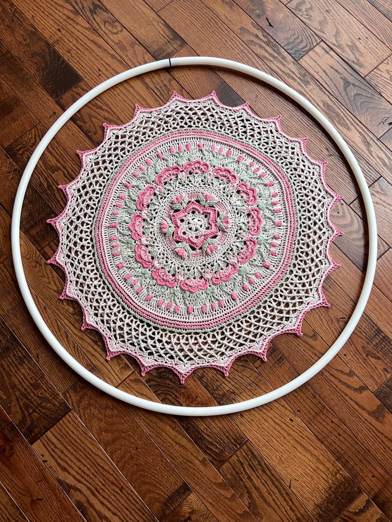 Pu&ograve; includere: Un mandala circolare all'uncinetto con intricati motivi floreali nelle tonalit&agrave; crema, rosa tenue e verde salvia. Il pezzo &egrave; montato all'interno di un grande cerchio in plastica bianca, che mette in mostra un design dettagliato con bordo smerlato.