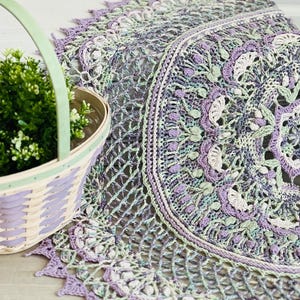 Pu&ograve; includere: Un centrino all'uncinetto rotondo e complesso con un motivo floreale a mandala nei toni della lavanda, del verde salvia e del crema. Accanto si trova un cesto intrecciato con un manico verde chiaro, contenente una piccola pianta verde.