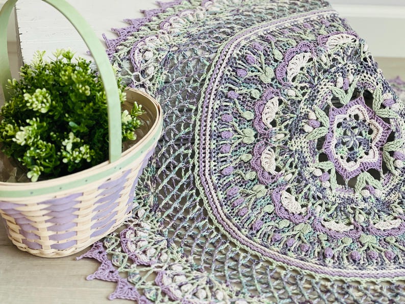 Pu&ograve; includere: Un centrino all'uncinetto rotondo e complesso con un motivo floreale a mandala nei toni della lavanda, del verde salvia e del crema. Accanto si trova un cesto intrecciato con un manico verde chiaro, contenente una piccola pianta verde.