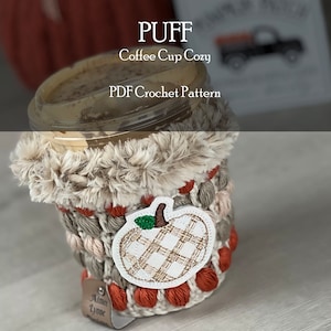 Puede incluir: Un calentador de taza de café tejido a crochet con un borde de piel sintética y una aplicación de calabaza. El calentador está en tonos beige, marrón y naranja. El texto "PUFF Coffee Cup Cozy PDF Crochet Pattern" es visible en la parte superior de la imagen.