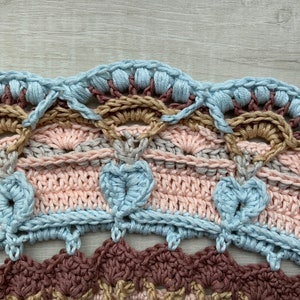 May include: Close-up of a crocheted doily with multiple layers of different colored yarn. The doily features a scalloped edge with a light blue color, followed by a brown, pink, and gray layer. The doily is crocheted in a circular pattern.