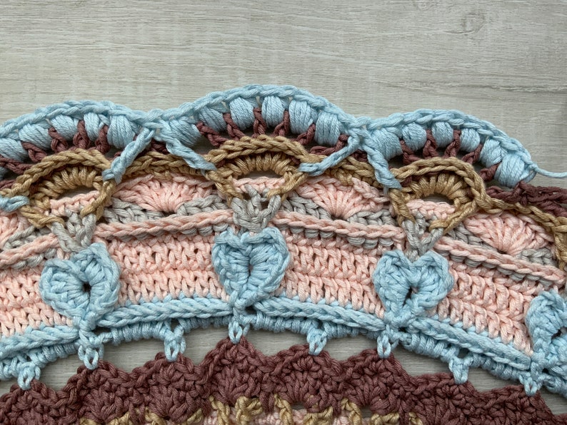May include: Close-up of a crocheted doily with multiple layers of different colored yarn. The doily features a scalloped edge with a light blue color, followed by a brown, pink, and gray layer. The doily is crocheted in a circular pattern.