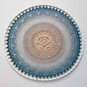 Pu&ograve; includere: Una decorazione da parete circolare con un dettagliato motivo a mandala all'uncinetto teso su un cerchio bianco. Il design sfuma da un centro beige chiaro a un gradiente di grigio e blu ottanio scuro sui bordi esterni con pizzo.