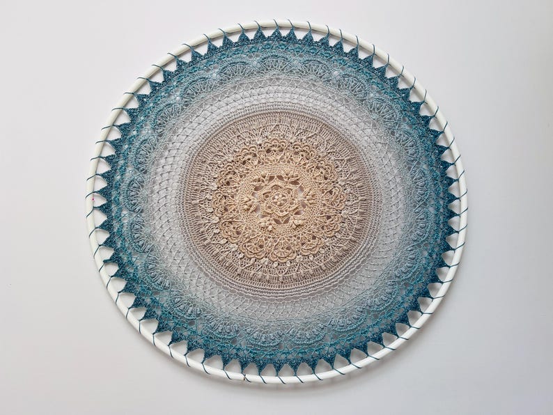 Pu&ograve; includere: Una decorazione da parete circolare con un dettagliato motivo a mandala all'uncinetto teso su un cerchio bianco. Il design sfuma da un centro beige chiaro a un gradiente di grigio e blu ottanio scuro sui bordi esterni con pizzo.