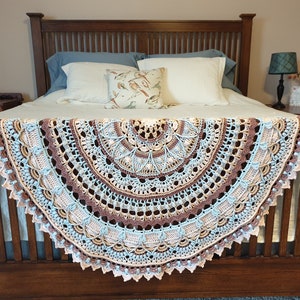 May include: A crocheted throw blanket in shades of brown, beige, blue, and pink. The blanket is draped over a wooden bed frame with white and blue pillows.