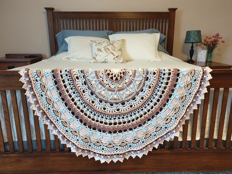 May include: A crocheted throw blanket in shades of brown, beige, blue, and pink. The blanket is draped over a wooden bed frame with white and blue pillows.