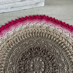 May include: A crocheted mandala with a pink and brown ombre design. The center of the mandala is a brown and beige pattern. The outer edge of the mandala is a solid pink.