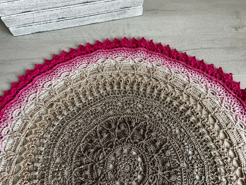 May include: A crocheted mandala with a pink and brown ombre design. The center of the mandala is a brown and beige pattern. The outer edge of the mandala is a solid pink.