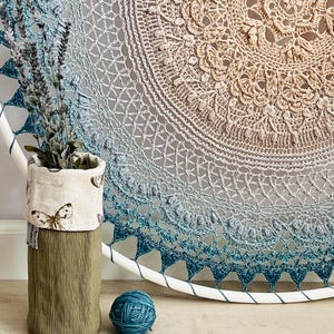 Pu&ograve; includere: Una grande decorazione murale circolare con un complesso mandala all'uncinetto in una sfumatura dal beige al blu profondo. Un contenitore cilindrico in tessuto con stampa a farfalle contiene lavanda essiccata, accanto a un gomitolo di lana blu.
