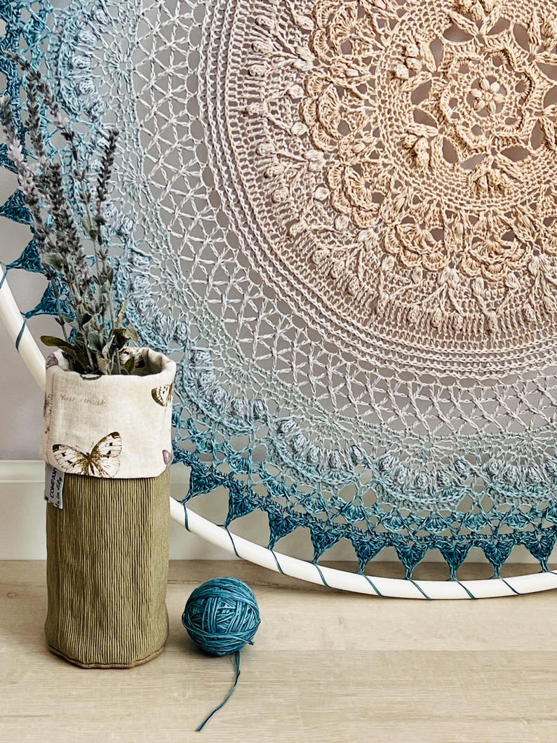 Pu&ograve; includere: Una grande decorazione murale circolare con un complesso mandala all'uncinetto in una sfumatura dal beige al blu profondo. Un contenitore cilindrico in tessuto con stampa a farfalle contiene lavanda essiccata, accanto a un gomitolo di lana blu.