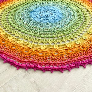 May include: A rainbow crochet mandala with a light blue center and a pink border. The mandala is made with a variety of colors, including red, orange, yellow, green, blue, and purple.