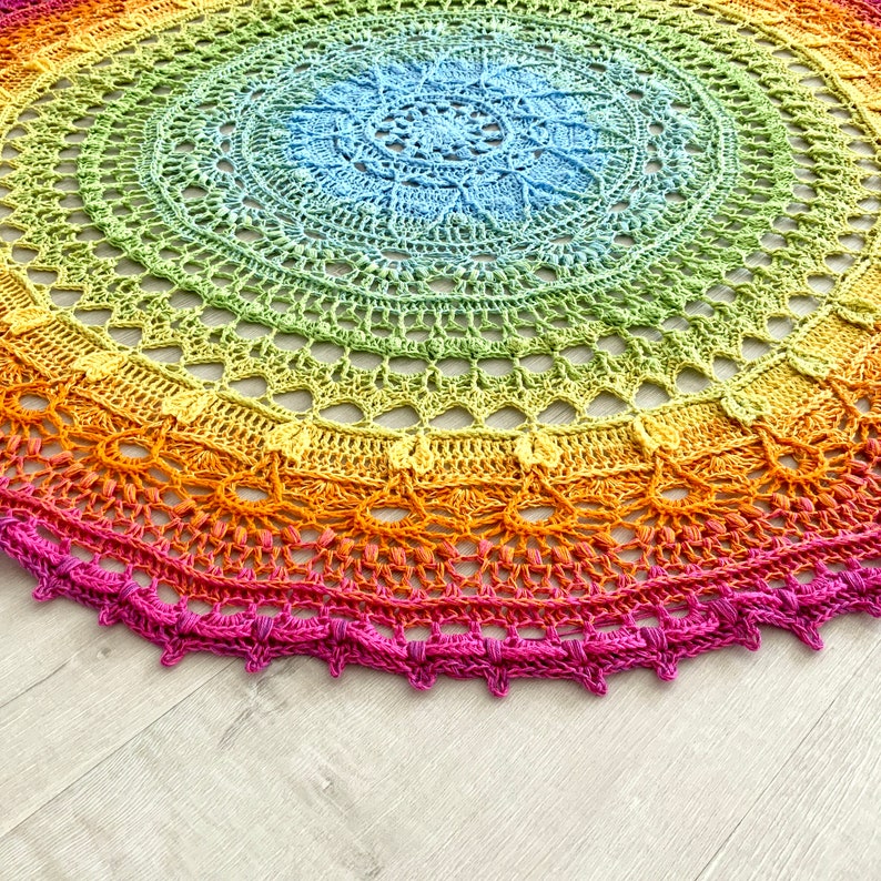 May include: A rainbow crochet mandala with a light blue center and a pink border. The mandala is made with a variety of colors, including red, orange, yellow, green, blue, and purple.