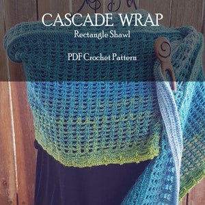 Può includere: Uno scialle all'uncinetto in tonalità di blu e verde, con un motivo a punto testurizzato. Lo scialle è appeso a un appendiabiti e ha una spilla per scialle in legno. Il testo "CASCADE WRAP" e "Rectangle Shawl" è visualizzato sopra lo scialle. Il testo "PDF Crochet Pattern" è visualizzato sotto lo scialle.