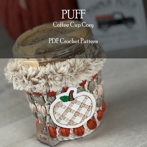 Puede incluir: Un calentador de taza de café tejido a crochet con un borde de piel sintética y una aplicación de calabaza. El calentador está en tonos beige, marrón y naranja. El texto "PUFF Coffee Cup Cozy PDF Crochet Pattern" es visible en la parte superior de la imagen.