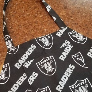 May include: Black and white kitchen apron with a repeating pattern of the Las Vegas Raiders logo. The logo features a silver helmet with crossed swords and the word "RAIDERS" written in a white, bold font.