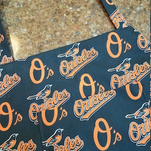 May include: Black apron with an orange and white repeating pattern of the Baltimore Orioles logo. The logo features a bird and the text "O's Orioles".