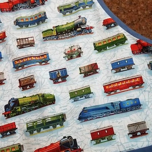 May include: A blue and white fabric apron with a train theme. The apron is covered in a repeating pattern of various trains, including red, green, and blue locomotives, boxcars, and tank cars. The apron has a blue trim and a large pocket.