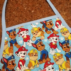 May include: A blue and white apron with a repeating pattern of cartoon dogs from the show Paw Patrol. The dogs are wearing their signature uniforms and are in various poses.