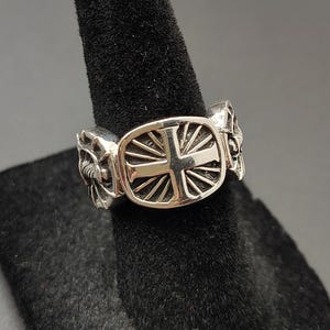 Könnte beinhalten: Ein silberfarbener Metallring mit einer zentralen rechteckigen Platte, die ein erhabenes Kreuzdesign umgeben von strahlenförmigen Linien zeigt. Die Ringschiene weist auf beiden Seiten detaillierte, skulpturale Verzierungen mit polierter Oberfläche auf.