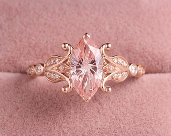 May include: A rose gold ring featuring a central marquise-cut peach-colored gemstone. The band is adorned with intricate metalwork and small clear stones on both sides, presented in a dusty pink velvet jewelry box with a square interior.