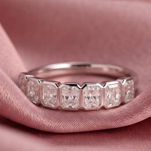 Puede incluir: Un anillo de banda plateada que presenta una serie de seis piedras rectangulares, transparentes y facetadas, engastadas en estilo bisel. Las piedras están dispuestas horizontalmente a lo largo de la mitad superior de la banda, creando un aspecto uniforme.