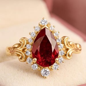 Könnte beinhalten: Ein Goldring mit einem großen birnenförmigen, tiefroten Edelstein in der Mitte, der von einem Kranz aus kleinen, runden, klaren Steinen umgeben ist. Die Ringschiene zeigt auf beiden Seiten kunstvolle Schnörkel, die von der Fassung ausgehen.